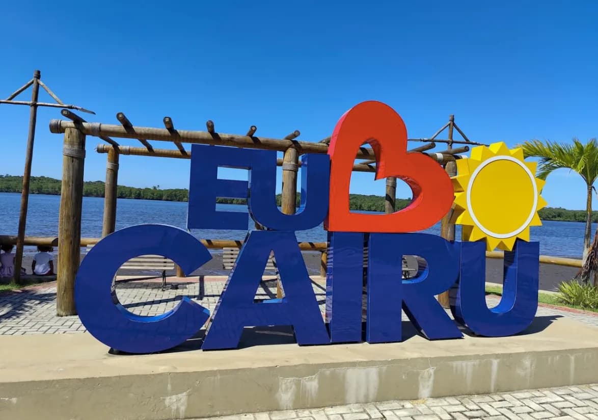 Letreiro 'Eu Amo Cairu' à beira-mar, com coqueiros e mangue ao fundo